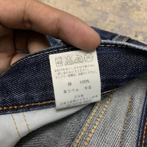 May include: Close-up of a dark blue denim fabric with visible stitching. A white tag with Japanese text and care symbols is attached. The tag includes the text "綿 100%" and "革ラベル 牛革".