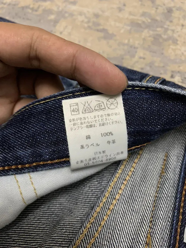 May include: Close-up of a dark blue denim fabric with visible stitching. A white tag with Japanese text and care symbols is attached. The tag includes the text "綿 100%" and "革ラベル 牛革".