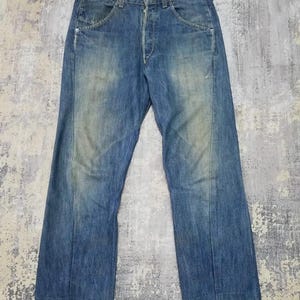 Puede incluir: Vaqueros de mezclilla azul descolorido con bragueta de botones y diseño clásico de cinco bolsillos. Los vaqueros muestran signos de desgaste y decoloración, dándoles un aspecto vintage. El corte recto y el lavado medio los convierten en un básico versátil.