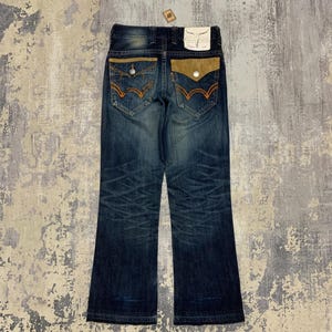 May include: Dark wash denim jeans with a bootcut leg. The back pockets have a unique design with tan suede-like fabric and orange stitching. A small tag is attached to the back waistband.