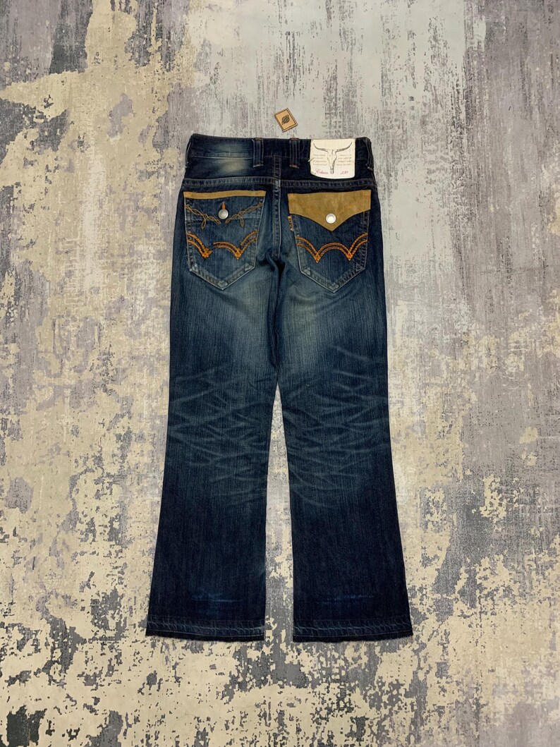 May include: Dark wash denim jeans with a bootcut leg. The back pockets have a unique design with tan suede-like fabric and orange stitching. A small tag is attached to the back waistband.