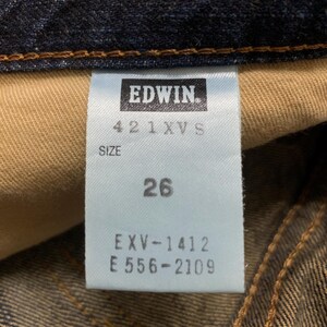 May include: Close-up of a blue denim tag with the brand name "EDWIN" in white. The tag also displays the size "26" and other product codes. The denim fabric is visible in the background.