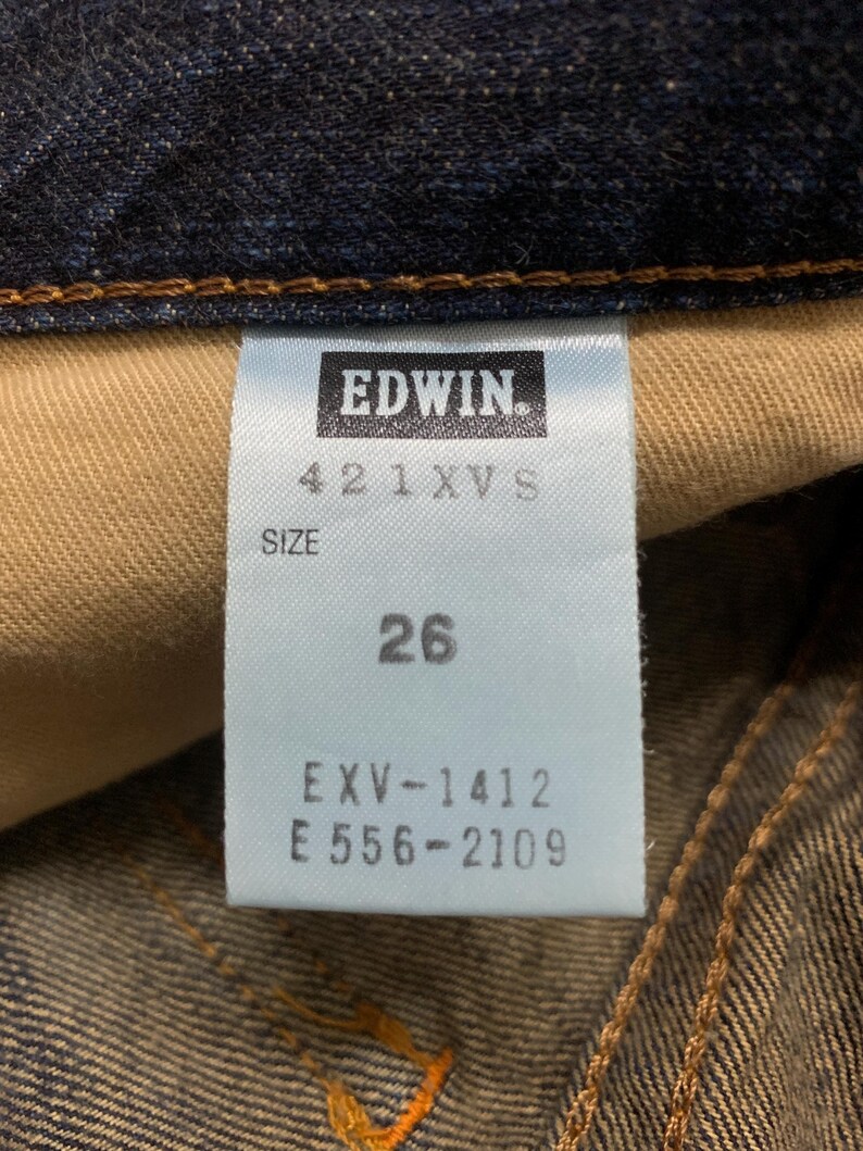 May include: Close-up of a blue denim tag with the brand name "EDWIN" in white. The tag also displays the size "26" and other product codes. The denim fabric is visible in the background.