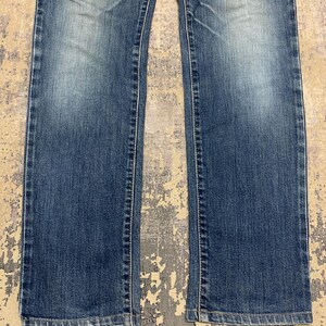 May include: Close-up of blue denim jeans. The jeans have a classic five-pocket design, with a straight leg cut. The denim has a faded wash, with lighter areas on the thighs and knees. White stitching is visible along the seams.