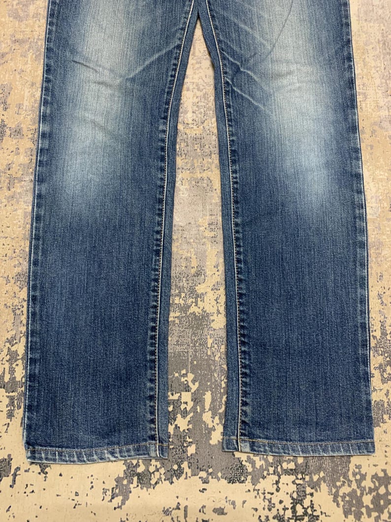 May include: Close-up of blue denim jeans. The jeans have a classic five-pocket design, with a straight leg cut. The denim has a faded wash, with lighter areas on the thighs and knees. White stitching is visible along the seams.
