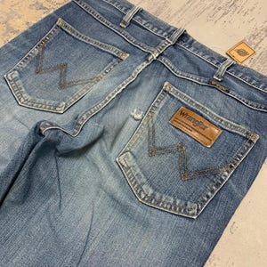 May include: Faded blue denim jeans with classic five-pocket styling. Features the Wrangler logo on a brown leather patch and signature stitching on the back pockets. A vintage tag is attached.