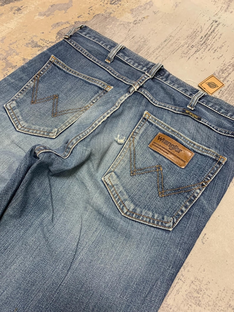 May include: Faded blue denim jeans with classic five-pocket styling. Features the Wrangler logo on a brown leather patch and signature stitching on the back pockets. A vintage tag is attached.