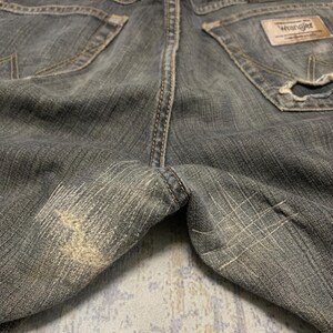 Pu&ograve; includere: Primo piano di jeans Wrangler grigi usurati. I pantaloni in denim mostrano segni di usura, con aree sbiadite e dettagli usurati. Le tasche posteriori e l'etichetta Wrangler sono visibili.