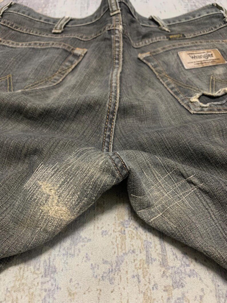Pu&ograve; includere: Primo piano di jeans Wrangler grigi usurati. I pantaloni in denim mostrano segni di usura, con aree sbiadite e dettagli usurati. Le tasche posteriori e l'etichetta Wrangler sono visibili.