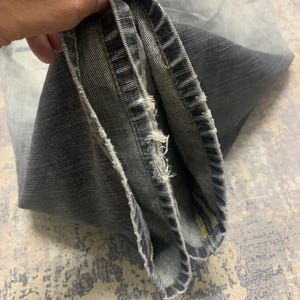 Pode incluir: Close-up de jeans cinza. Os jeans t&ecirc;m uma lavagem desbotada e detalhes desgastados ao longo da bainha. O tecido est&aacute; dobrado, revelando a constru&ccedil;&atilde;o interna e as costuras. O visual geral &eacute; casual e usado.