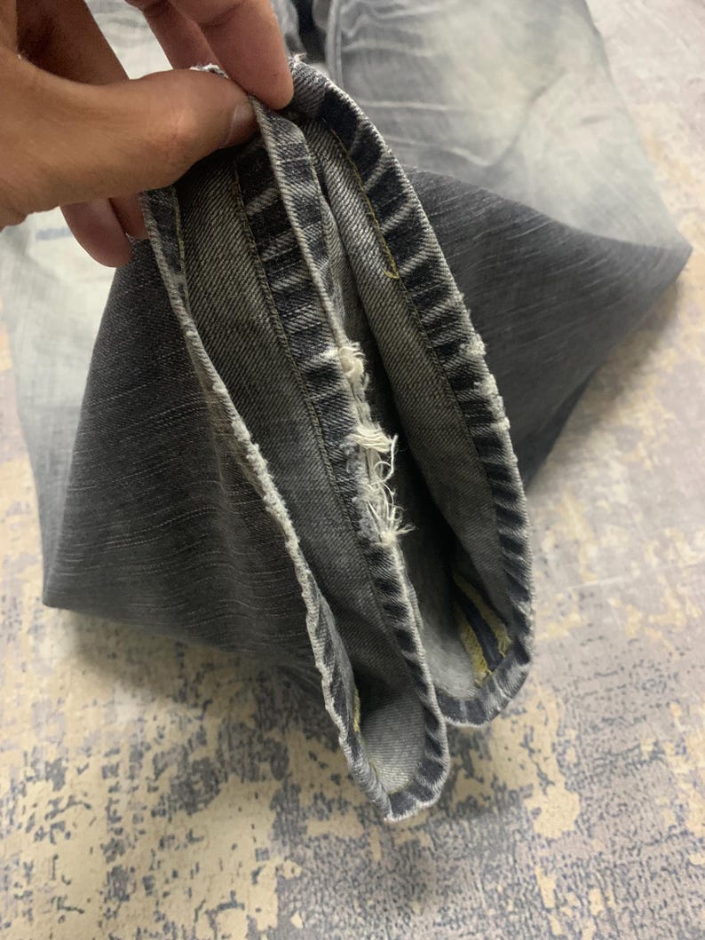 Pode incluir: Close-up de jeans cinza. Os jeans t&ecirc;m uma lavagem desbotada e detalhes desgastados ao longo da bainha. O tecido est&aacute; dobrado, revelando a constru&ccedil;&atilde;o interna e as costuras. O visual geral &eacute; casual e usado.