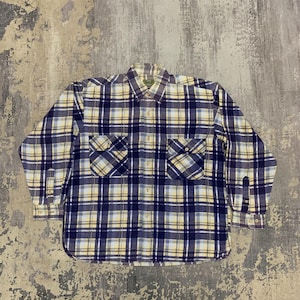 Può includere: Una camicia a maniche lunghe con bottoni e motivo a quadri blu navy, giallo e bianco. La camicia ha due tasche sul petto e polsini con bottoni. È appoggiata su una superficie grigia strutturata.