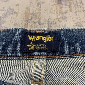 May include: Close-up of a Wrangler jeans label. The navy blue label features the brand name in yellow, with a star and text that reads "CRAFTED WITH PRIDE". The denim fabric is visible, with detailed stitching.