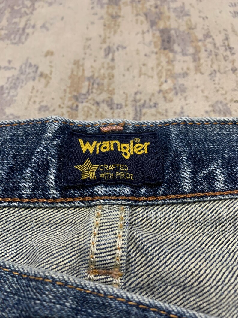May include: Close-up of a Wrangler jeans label. The navy blue label features the brand name in yellow, with a star and text that reads "CRAFTED WITH PRIDE". The denim fabric is visible, with detailed stitching.