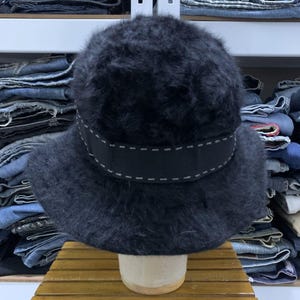 May include: A black, fuzzy hat with a wide, dark ribbon band featuring white stitching. The hat has a rounded crown and a brim. The hat is displayed on a wooden stand.