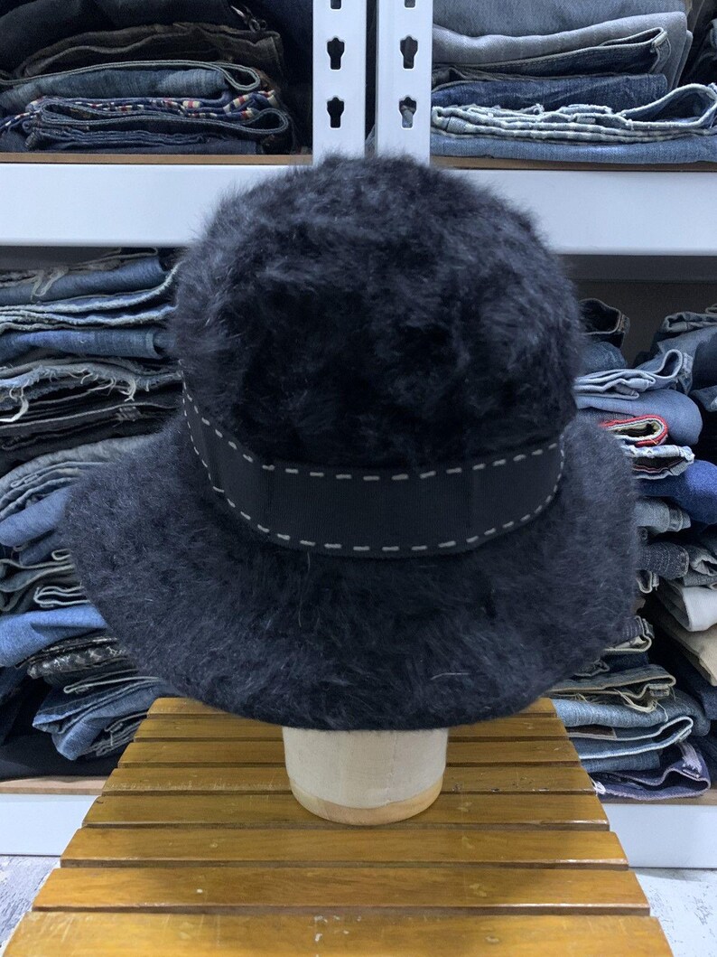 May include: A black, fuzzy hat with a wide, dark ribbon band featuring white stitching. The hat has a rounded crown and a brim. The hat is displayed on a wooden stand.