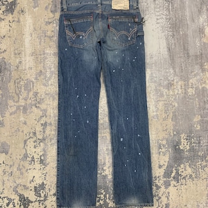 May include: Faded blue denim jeans with white paint splatters. The jeans have a classic five-pocket design, red stitching details, and a "BLUE TRIP" label. The back pockets have a distinctive curved stitch.