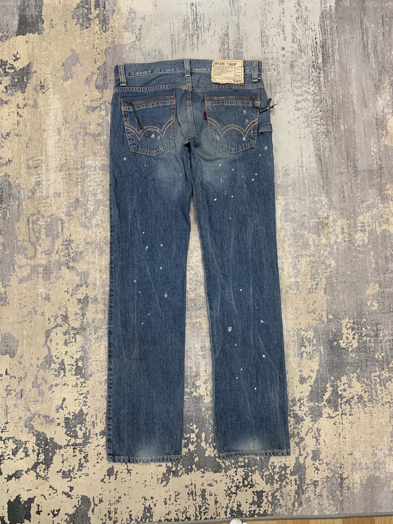 May include: Faded blue denim jeans with white paint splatters. The jeans have a classic five-pocket design, red stitching details, and a "BLUE TRIP" label. The back pockets have a distinctive curved stitch.