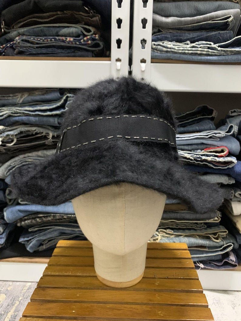 May include: A black, fuzzy hat with a wide brim and a black ribbon band. The band is accented with white stitching. The hat is displayed on a mannequin head.