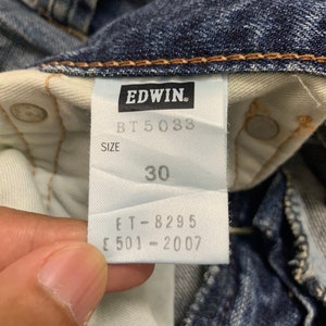 May include: Close-up of a pair of blue denim jeans with a white tag. The tag features the brand name "EDWIN" and the size "30". Additional text includes "BT 5033", "ET-8295", and "E 501-2007".