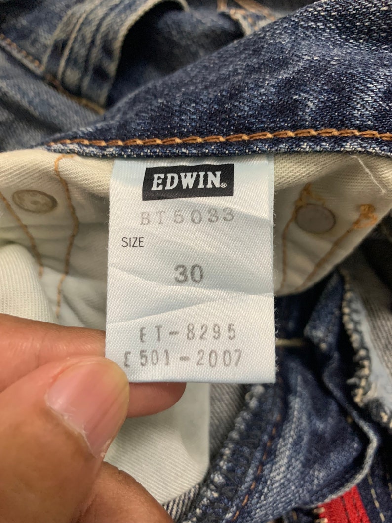 May include: Close-up of a pair of blue denim jeans with a white tag. The tag features the brand name "EDWIN" and the size "30". Additional text includes "BT 5033", "ET-8295", and "E 501-2007".