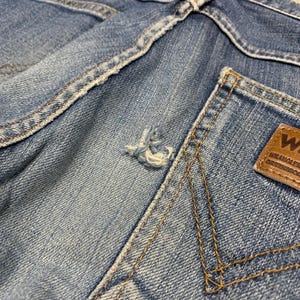 May include: Close-up of light blue denim jeans, showing the back pocket and waistband. The jeans have a distressed look with a small tear and visible stitching. A brown leather patch with the brand logo is on the back pocket.