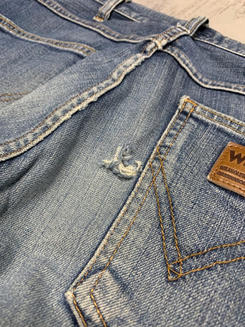 May include: Close-up of light blue denim jeans, showing the back pocket and waistband. The jeans have a distressed look with a small tear and visible stitching. A brown leather patch with the brand logo is on the back pocket.
