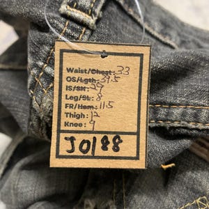 Pu&ograve; includere: Primo piano di un paio di jeans neri sbiaditi. Un'etichetta attaccata ai jeans mostra le misure: Vita/Petto 84 cm, Lunghezza totale 100 cm, Interno gamba 74 cm, Gamba/Orlo 20 cm, Anteriore/Orlo 29 cm, Coscia 30 cm, Ginocchio 23 cm. L'etichetta ha anche il numero "J0188".