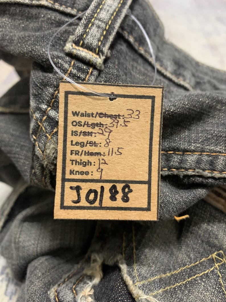Pu&ograve; includere: Primo piano di un paio di jeans neri sbiaditi. Un'etichetta attaccata ai jeans mostra le misure: Vita/Petto 84 cm, Lunghezza totale 100 cm, Interno gamba 74 cm, Gamba/Orlo 20 cm, Anteriore/Orlo 29 cm, Coscia 30 cm, Ginocchio 23 cm. L'etichetta ha anche il numero "J0188".