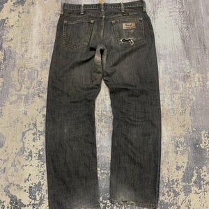 Pu&ograve; includere: Jeans in denim blu scuro con un aspetto usurato. I jeans presentano un classico design a cinque tasche, una toppa Wrangler sulla tasca posteriore e un'area usurata sulla tasca posteriore destra. I jeans sono stesi, mostrando la vista posteriore.