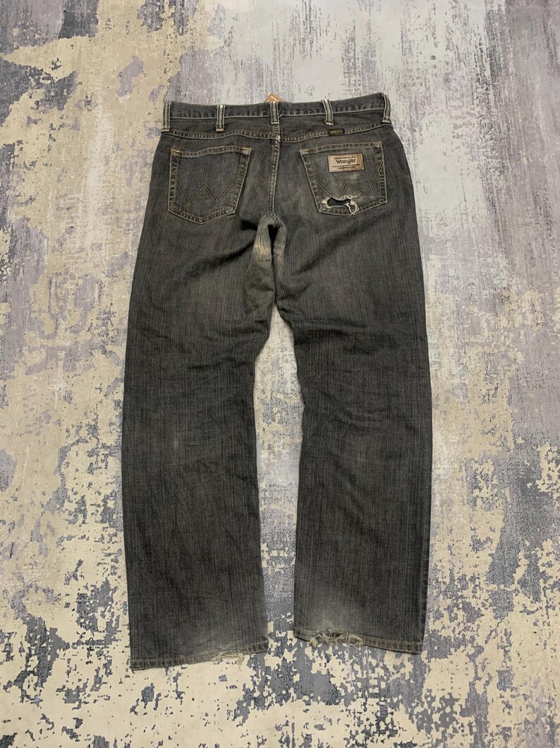 Pu&ograve; includere: Jeans in denim blu scuro con un aspetto usurato. I jeans presentano un classico design a cinque tasche, una toppa Wrangler sulla tasca posteriore e un'area usurata sulla tasca posteriore destra. I jeans sono stesi, mostrando la vista posteriore.