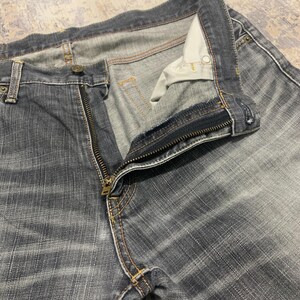 Pode incluir: Close-up de um par de jeans de ganga pretos desbotados com um z&iacute;per de lat&atilde;o parcialmente aberto. Os jeans t&ecirc;m um design cl&aacute;ssico de cinco bolsos com costuras vis&iacute;veis e fecho de bot&atilde;o. O tecido mostra um padr&atilde;o listrado sutil.