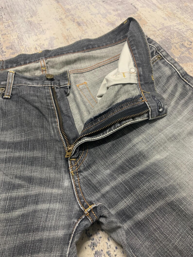 Pode incluir: Close-up de um par de jeans de ganga pretos desbotados com um z&iacute;per de lat&atilde;o parcialmente aberto. Os jeans t&ecirc;m um design cl&aacute;ssico de cinco bolsos com costuras vis&iacute;veis e fecho de bot&atilde;o. O tecido mostra um padr&atilde;o listrado sutil.