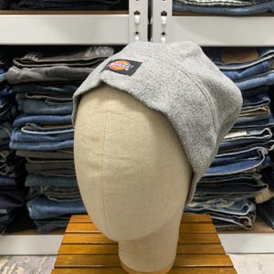 May include: A light gray fleece hat with a small rectangular patch featuring the Dickies logo in red, orange, and black. The hat is displayed on a mannequin head. The background shows shelves of folded denim jeans.
