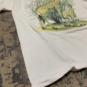 Puede incluir: Camiseta blanca con una ilustraci&oacute;n detallada de dos jirafas y un &aacute;rbol. Las jirafas tienen abrigos estampados en marr&oacute;n y amarillo, de pie sobre hierba verde. La camiseta est&aacute; hecha de tela blanca suave.