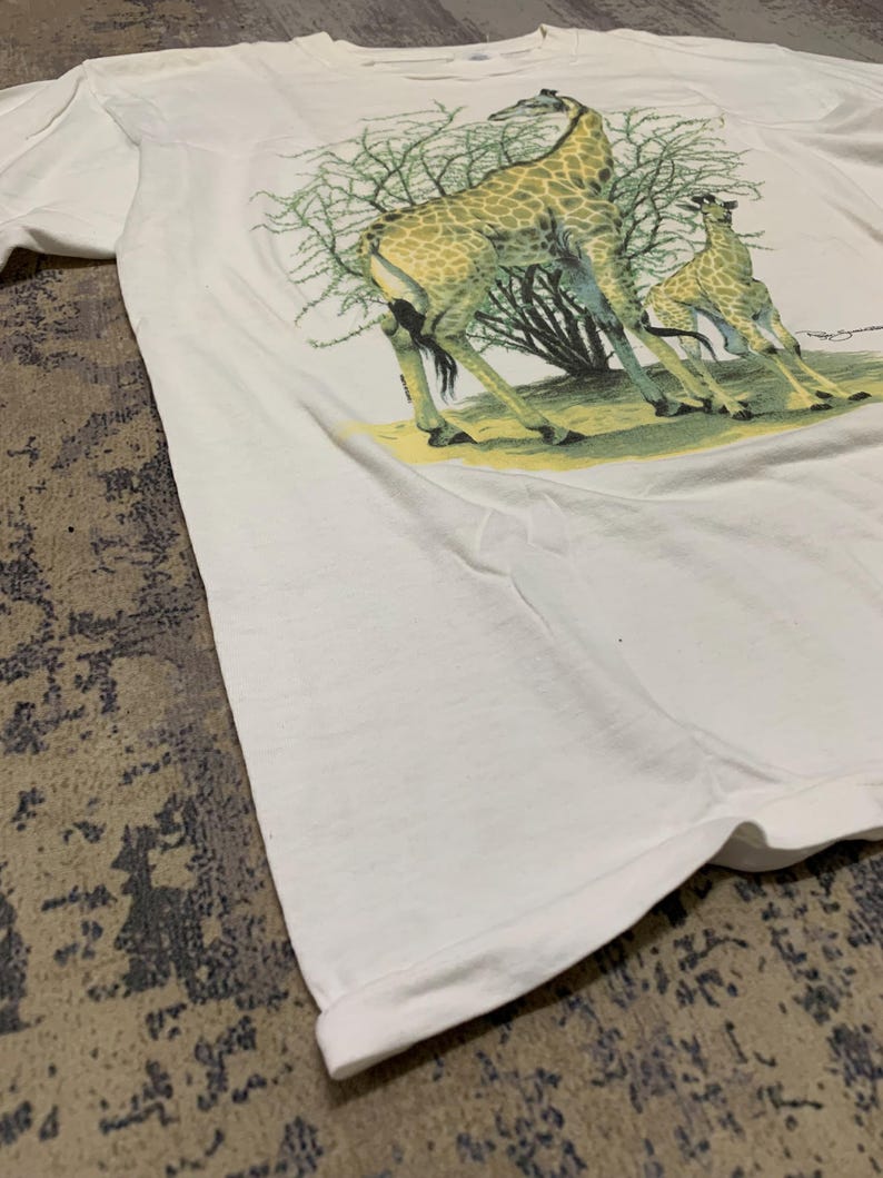Puede incluir: Camiseta blanca con una ilustraci&oacute;n detallada de dos jirafas y un &aacute;rbol. Las jirafas tienen abrigos estampados en marr&oacute;n y amarillo, de pie sobre hierba verde. La camiseta est&aacute; hecha de tela blanca suave.