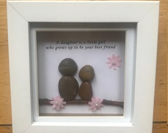 Puede incluir: Una pieza de arte de guijarros enmarcada con dos guijarros marrones que representan a un padre y un hijo sentados en una rama con flores rosas. La cita "Una hija es una niña pequeña que crece para ser tu mejor amiga" está escrita en el fondo blanco.
