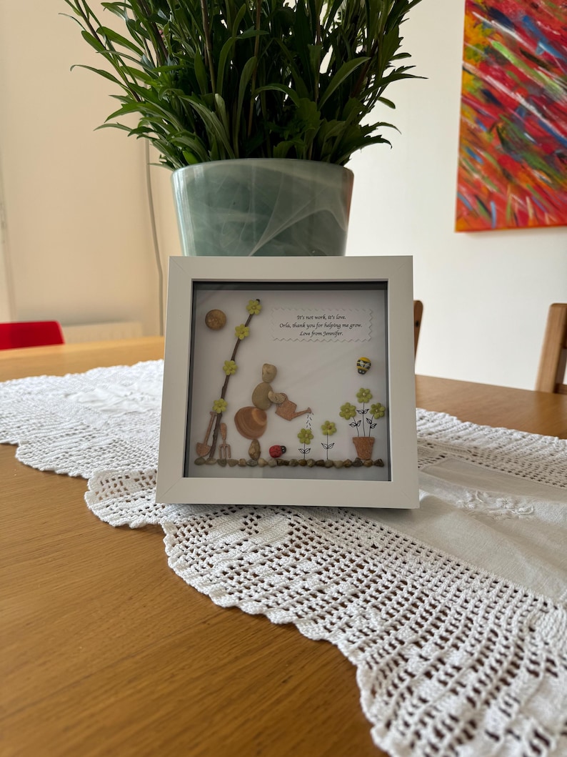 Puede incluir: Una imagen enmarcada en blanco con un dise&ntilde;o de arte de guijarros de un oso regando flores. El texto de la imagen dice "No es trabajo, es amor.  Oh, gracias por ayudarme a crecer. Amor de [Nombre]"