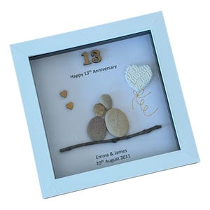 May include: A white framed picture with a white background. The picture features a pebble art design of a couple holding hands, a heart shaped balloon, and the number 13. The text reads "Happy 13th Anniversary, Emma & James, 20th August 2011".