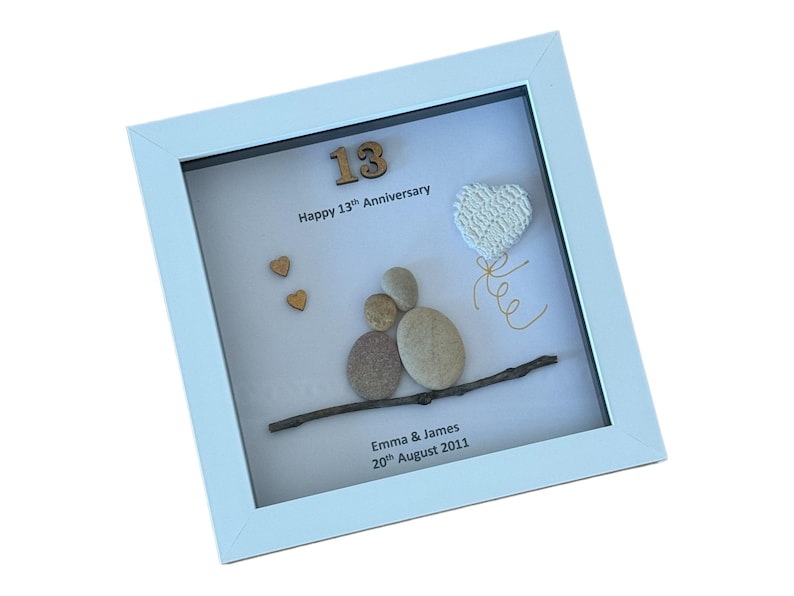 May include: A white framed picture with a white background. The picture features a pebble art design of a couple holding hands, a heart shaped balloon, and the number 13. The text reads "Happy 13th Anniversary, Emma & James, 20th August 2011".