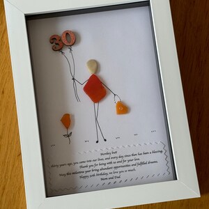 May include: A framed piece of art with a hand-drawn figure holding a heart-shaped object and a balloon with the number 30 on it. The art is made with colorful sea glass and the frame is white. The text below the art reads "Monkey butt thirty years ago, you came into our lives, and every day since then has been a blessing. Thank you for being with us and for your love. May this milestone year bring abundant opportunities and fulfilled dreams. Happy 30th birthday, we love you so much. Mom and Dad."