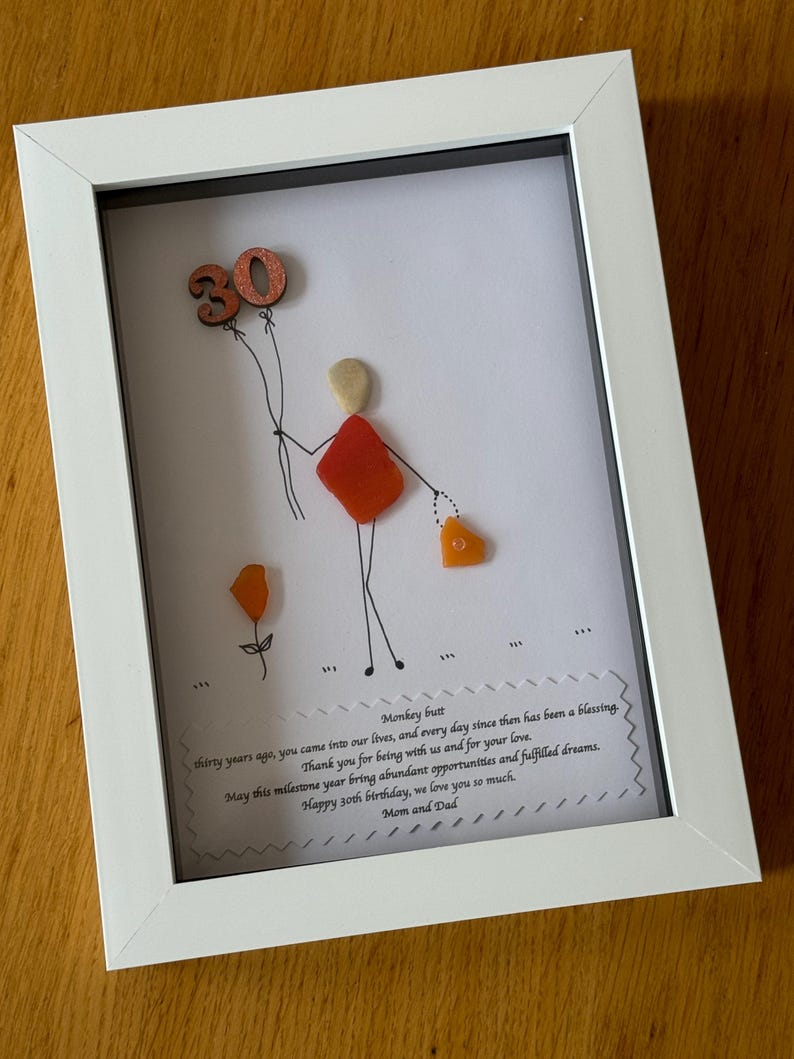 May include: A framed piece of art with a hand-drawn figure holding a heart-shaped object and a balloon with the number 30 on it. The art is made with colorful sea glass and the frame is white. The text below the art reads "Monkey butt thirty years ago, you came into our lives, and every day since then has been a blessing. Thank you for being with us and for your love. May this milestone year bring abundant opportunities and fulfilled dreams. Happy 30th birthday, we love you so much. Mom and Dad."