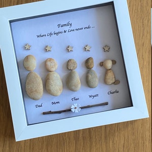 May include: A framed family artwork in a white frame. Pebble figures represent family members, with wooden stars above. The names "Dad", "Mom", "Theo", "Wyatt", and "Charlie" are written below. The text "Family Where Life begins & Love never ends..." is also included.
