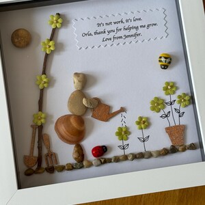 Puede incluir: Una obra de arte enmarcada que presenta a una persona de guijarros regando flores con una regadera. La obra de arte est&aacute; hecha con materiales naturales como guijarros, conchas y ramitas. El fondo es blanco con un mensaje escrito a mano que dice "No es trabajo, es amor. Orla, gracias por ayudarme a crecer. Con amor, Jennifer."