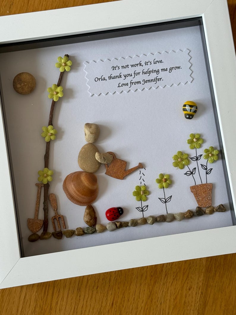 Puede incluir: Una obra de arte enmarcada que presenta a una persona de guijarros regando flores con una regadera. La obra de arte est&aacute; hecha con materiales naturales como guijarros, conchas y ramitas. El fondo es blanco con un mensaje escrito a mano que dice "No es trabajo, es amor. Orla, gracias por ayudarme a crecer. Con amor, Jennifer."