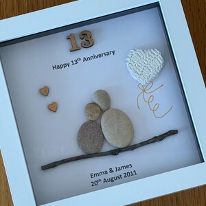 May include: A white frame with a white background. The number 13 is made of wood and is in the top left corner. The text "Happy 13th Anniversary" is below the number. There are two small wooden hearts in the top left corner. A white crocheted heart is in the top right corner. A twig with five gray stones stacked on top of it is in the center of the frame. The text "Emma & James 20th August 2011" is at the bottom of the frame.
