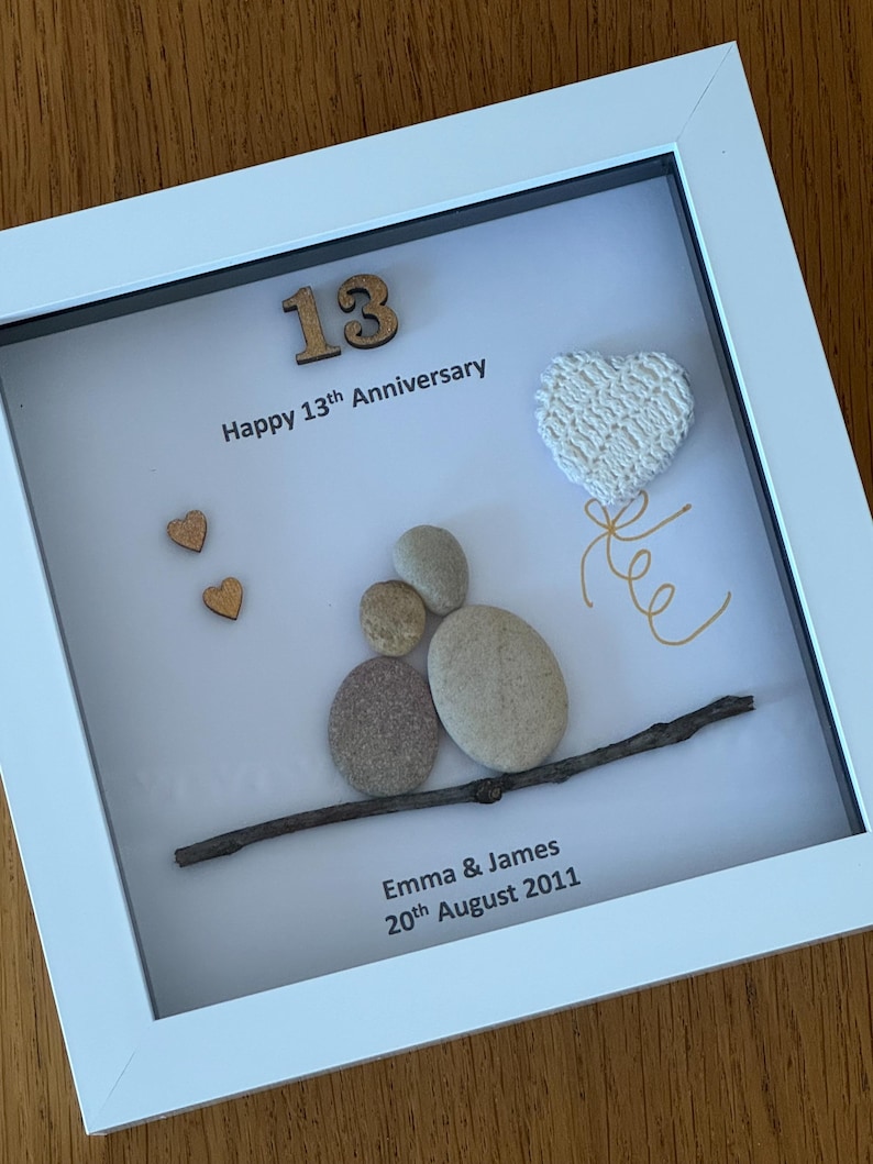 May include: A white frame with a white background. The number 13 is made of wood and is in the top left corner. The text "Happy 13th Anniversary" is below the number. There are two small wooden hearts in the top left corner. A white crocheted heart is in the top right corner. A twig with five gray stones stacked on top of it is in the center of the frame. The text "Emma & James 20th August 2011" is at the bottom of the frame.