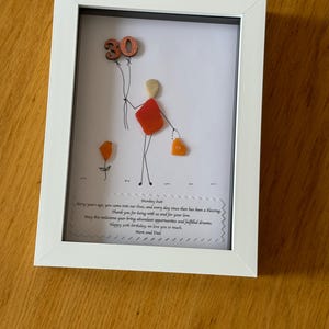 May include: A framed piece of art made with colorful stones and beads. The art depicts a person holding balloons with the number 30 on them. The frame has a white mat and a white frame. The text in the frame reads "Monkey butt thirty years ago, you came into our lives, and every day since then has been a blessing Thank you for being with us and for your love. May this milestone year bring abundant opportunities and fulfilled dreams. Happy 30th birthday, we love you so much. Mom and Dad"