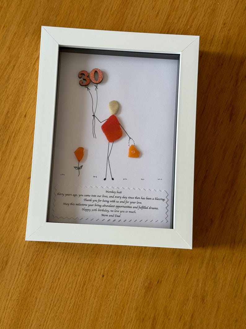 May include: A framed piece of art made with colorful stones and beads. The art depicts a person holding balloons with the number 30 on them. The frame has a white mat and a white frame. The text in the frame reads "Monkey butt thirty years ago, you came into our lives, and every day since then has been a blessing Thank you for being with us and for your love. May this milestone year bring abundant opportunities and fulfilled dreams. Happy 30th birthday, we love you so much. Mom and Dad"