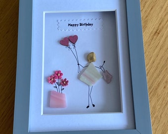May include: A framed artwork featuring a hand-drawn figure holding pink heart balloons and a pink gift bag. The figure is made of colorful stones and is surrounded by pink flowers in a pink pot. The frame is gray and the background is white with the text "Happy Birthday" in a white banner.